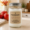 A glass mason jar candle with a silver metal lid and twine detail, labeled 'Country Crackle Candle' in 'Apple Orchard' scented wax, set on a kitchen counter with red apples in the background.