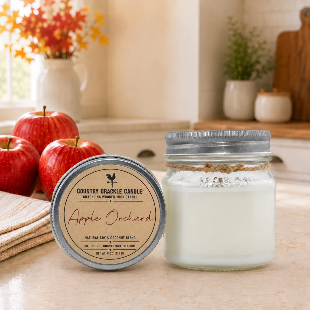 A Country Crinkle Candle in an Apple Orchard scent, shown as a white wax candle in a glass jar with a silver lid next to its matching tin container. The scene is set on a kitchen counter with decorative red apples and a blurred kitchen background.