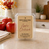 A clear plastic clamshell package of Country Crackle Candle soy wax melts labeled 'Apple Orchard' from the Fall Collection, set on a kitchen counter with red apples and a blurred kitchen background.