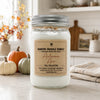 A white soy wax candle in a clear glass mason jar with a silver metal lid and a kraft paper label reading 'Country Crackle Candle Autumn Air Fall Collection.' The jar sits on a light countertop in a styled home setting with small pumpkins, a folded towel, and warm background decor.