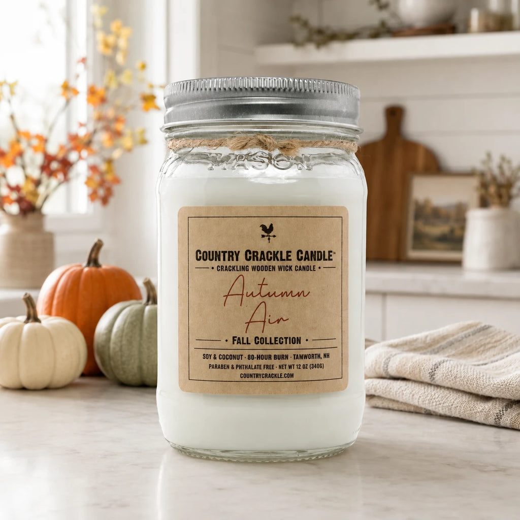 A white soy wax candle in a clear glass mason jar with a silver metal lid and a kraft paper label reading 'Country Crackle Candle Autumn Air Fall Collection.' The jar sits on a light countertop in a styled home setting with small pumpkins, a folded towel, and warm background decor.