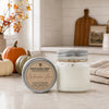 A jar candle and its tin lid are displayed on a kitchen counter with fall-themed props including pumpkins, a cutting board, and a striped towel. The label reads 'Country Crackle Candle' and 'Autumn Air'.