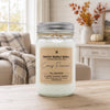 A glass jar candle with a silver metal lid and a beige label reading 'Country Crackle Candle Cozy Flannel' sits on a wooden table in a softly lit living room setting.
