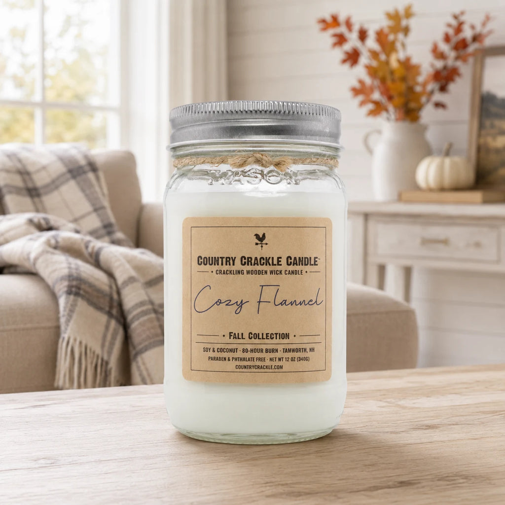 A glass jar candle with a silver metal lid and a beige label reading 'Country Crackle Candle Cozy Flannel' sits on a wooden table in a softly lit living room setting.