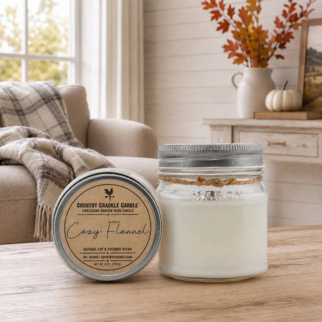Two jar candles on a wooden table in a cozy living room setting. One candle is open, showing white wax inside a clear glass jar with a silver metal lid and twine detail.