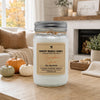 A jar candle labeled 'Country Crackle Candle Pumpkin Spice Fall Collection' sits on a wooden table in a cozy living room setting with pumpkins, a fireplace, and a couch in the background.