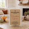 A clear plastic clamshell package of Country Crackle Candle 'Pumpkin Spice' soy wax melts sits on a wooden table in a cozy living room setting with pumpkins, a fireplace, and a couch in the background.