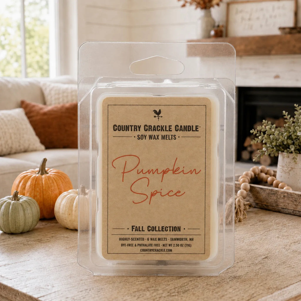 A clear plastic clamshell package of Country Crackle Candle 'Pumpkin Spice' soy wax melts sits on a wooden table in a cozy living room setting with pumpkins, a fireplace, and a couch in the background.