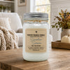 A glass jar candle with a silver metal lid sits on a rustic wooden table in a cozy living room setting. The candle has a beige label that reads 'Country Crackle Candle Sweater Season Fall Collection'.