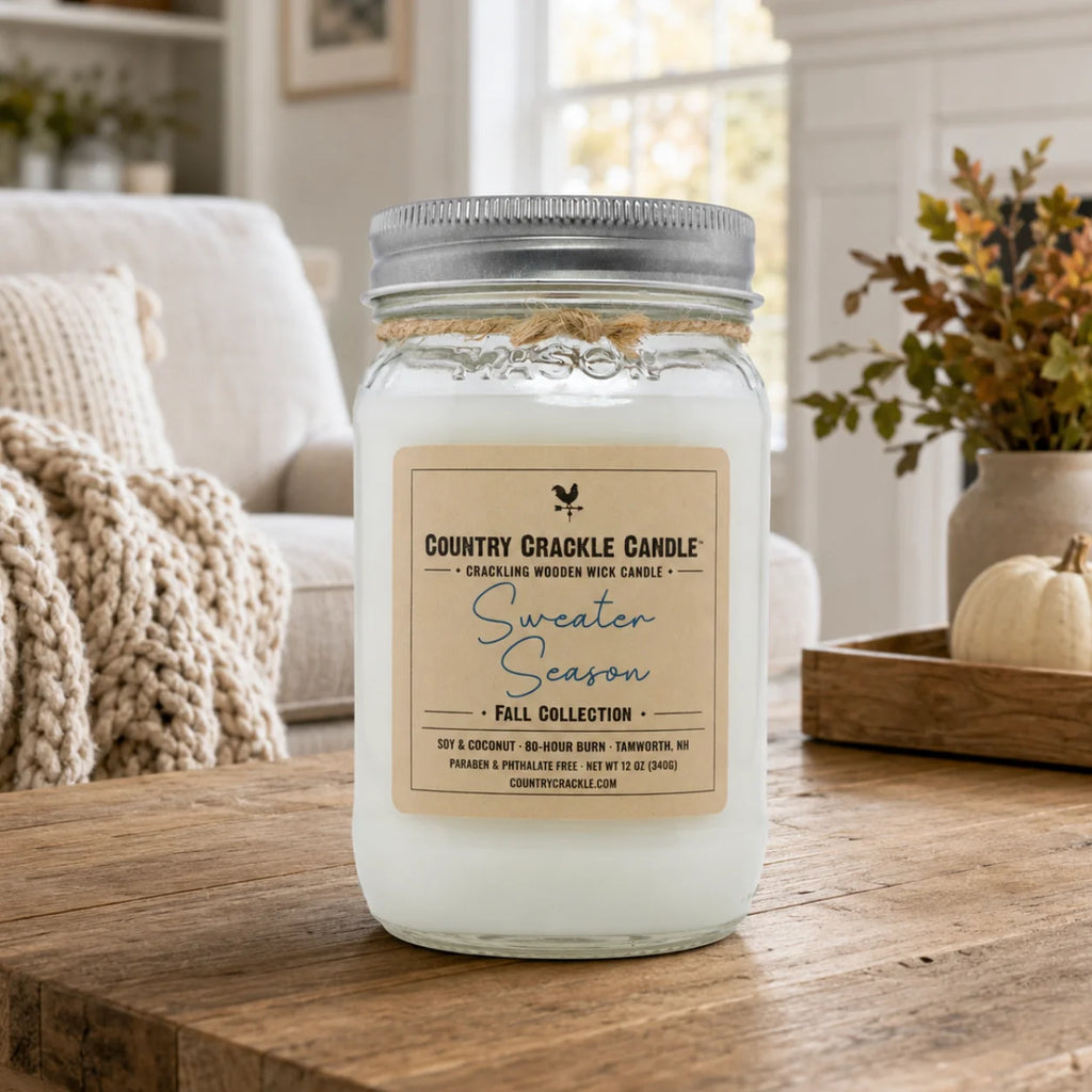 A glass jar candle with a silver metal lid sits on a rustic wooden table in a cozy living room setting. The candle has a beige label that reads 'Country Crackle Candle Sweater Season Fall Collection'.