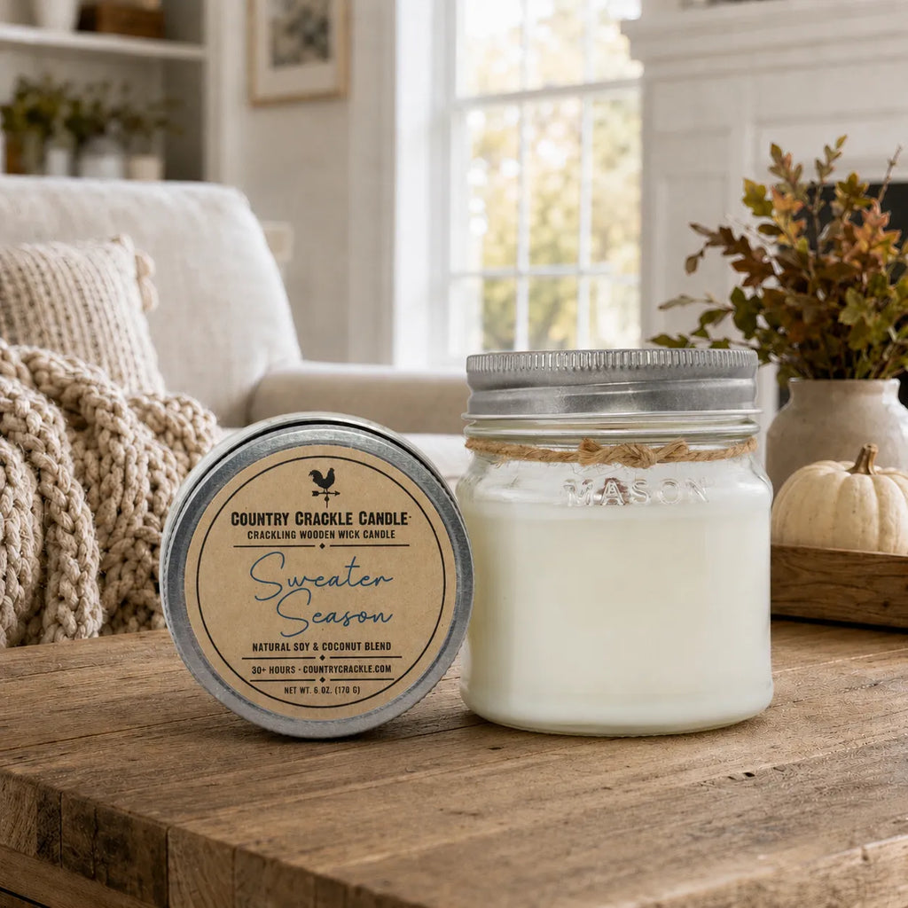 A jar candle and its lid are displayed on a rustic wooden table in a cozy living room setting. The candle is in a clear glass jar with white wax and a silver metal lid, while the lid features a round label that reads 'Country Crackle Candle' and 'Sweater Season'.