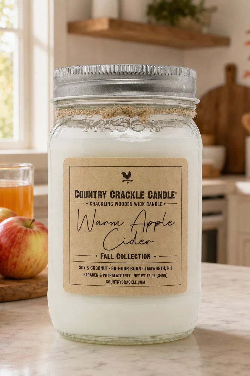 A large glass jar candle with a silver metal lid and a beige label reading 'Country Crackle Candle' and 'Warm Apple Cider' stands on a kitchen counter. The background shows a softly blurred kitchen scene with apples, a mug of orange drink, and warm lighting.