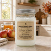 A large glass jar candle with a silver metal lid and a beige label reading 'Country Crackle Candle' and 'Warm Apple Cider' stands on a kitchen counter. The background shows a softly blurred kitchen scene with apples, a mug of orange drink, and warm lighting.