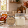 A mason jar candle with a silver lid and twine band stands on a kitchen counter beside its round lid label, which reads 'Country Crackle Candle Warm Apple Cider.' Red apples, a glass mug of amber liquid, and a softly blurred kitchen interior are visible in the background.