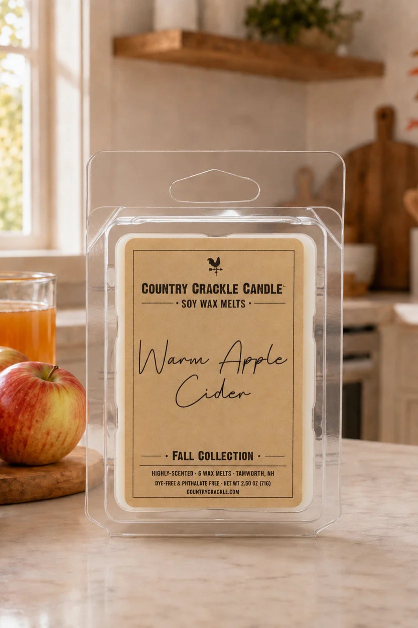 A packaged soy wax melt labeled 'Warm Apple Cider' by Country Crinkle Candle, displayed on a kitchen counter with apples and a glass of apple cider nearby.