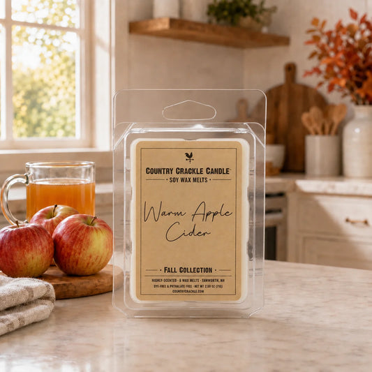 A packaged soy wax melt labeled 'Warm Apple Cider' by Country Crinkle Candle, displayed on a kitchen counter with apples and a glass of apple cider nearby.