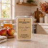 A packaged soy wax melt labeled 'Warm Apple Cider' by Country Crinkle Candle, displayed on a kitchen counter with apples and a glass of apple cider nearby.